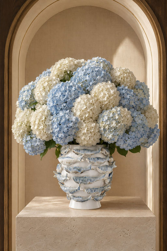 Vase Poissons Blancs en céramique blanche avec poissons en relief bleu-gris, présenté avec un bouquet d'hortensias blancs et bleus dans une niche en pierre, DUNE & CO